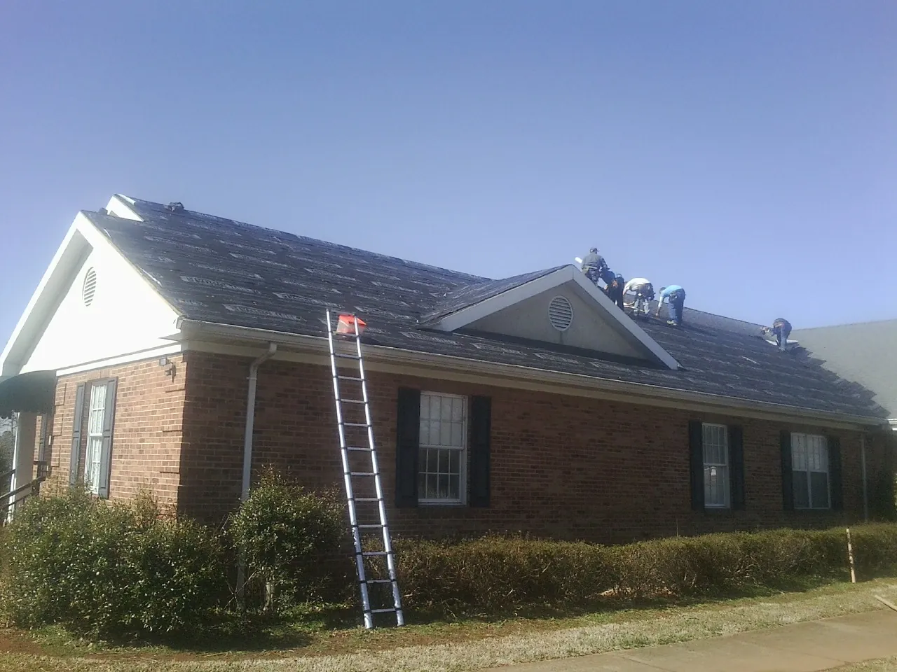 Crew tearing shingles off a brick ranch roof with a ladder set — Metal Roof Repair work by our crew in Junction