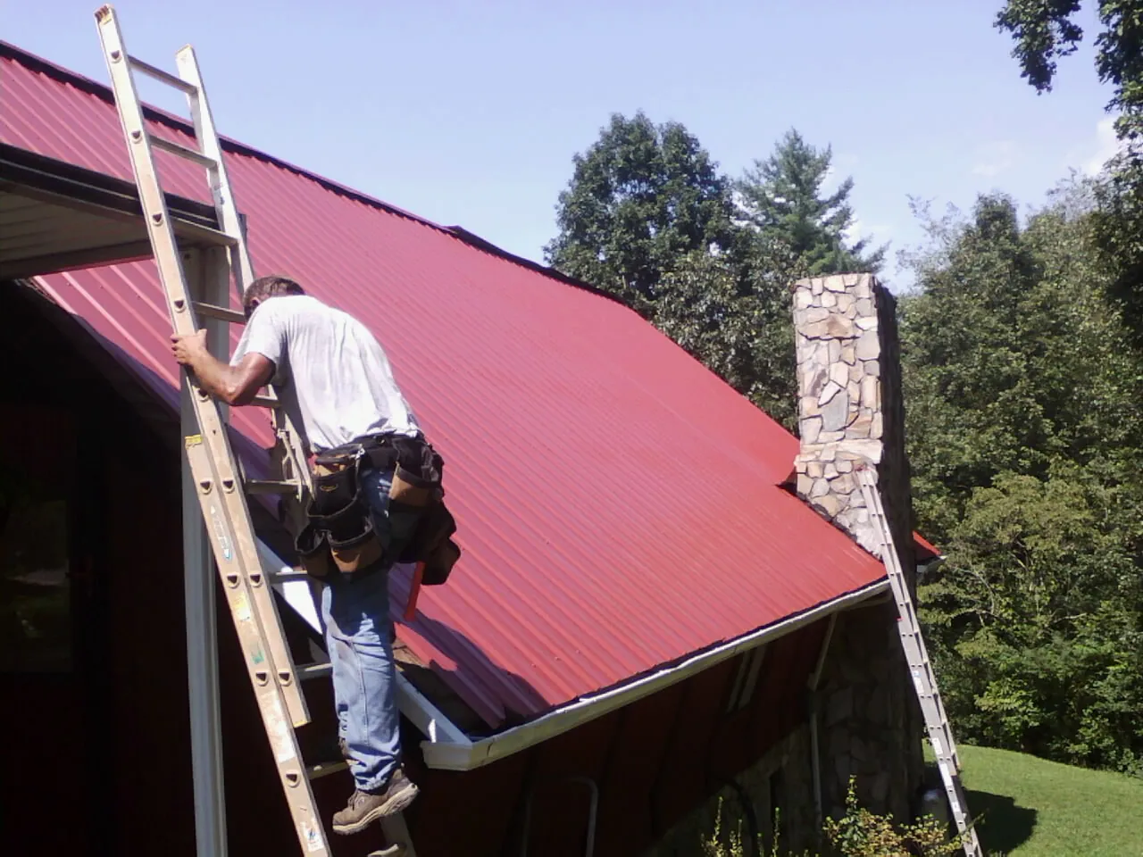 Roofer on a ladder beside a red metal roof and stone chimney — Soffit & Fascia Repair by Roofing Brothers in Junction